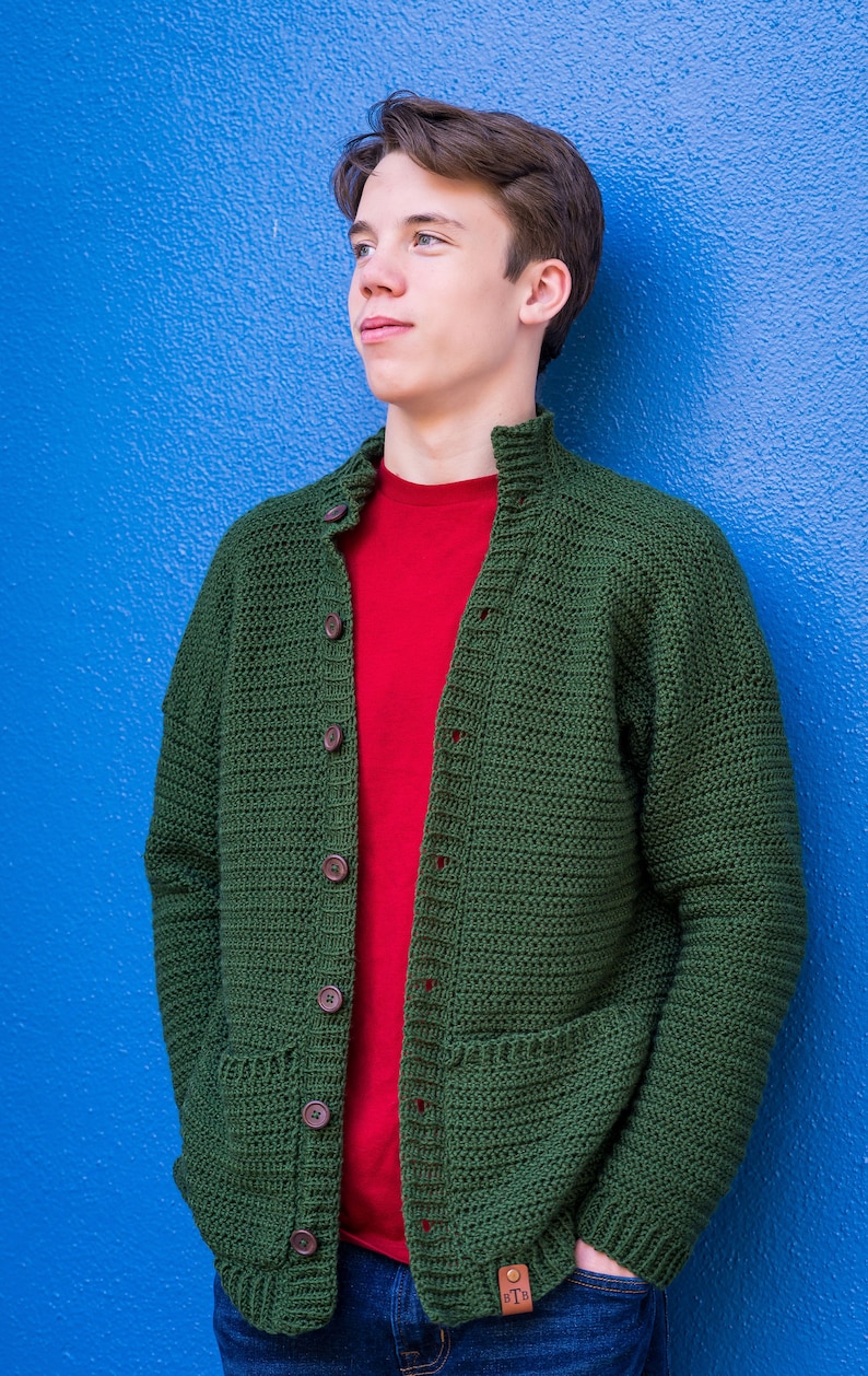 May include: A green crocheted cardigan with a red t-shirt underneath and blue jeans. The cardigan has a button closure and two pockets.