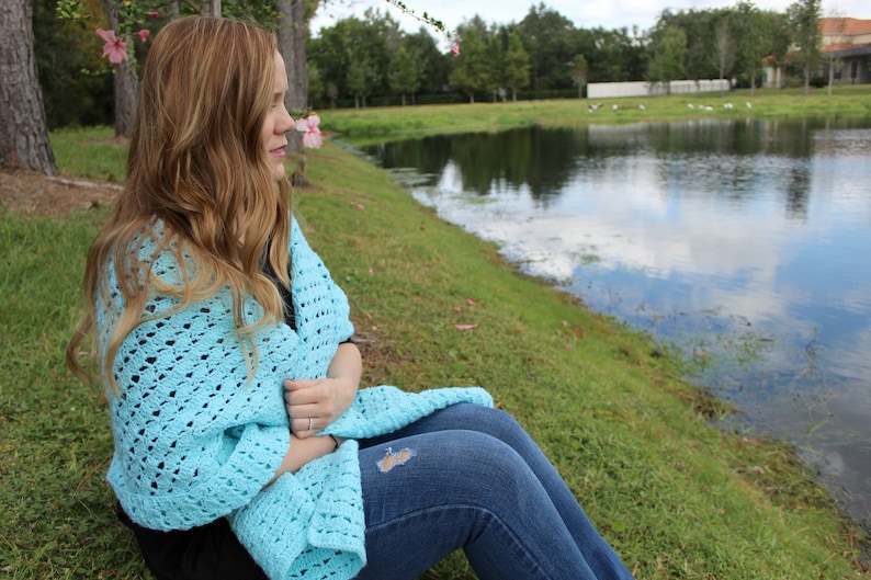 May include: A light blue crocheted shawl draped over the shoulders. The open weave design is visible. The person is wearing blue jeans and sitting on a grassy bank next to a pond. The background includes trees and a body of water.