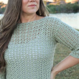 May include: A light green crochet sweater with three-quarter sleeves. The open weave design is visible, and the sweater has a boat neck. The garment is made of a soft, textured yarn.