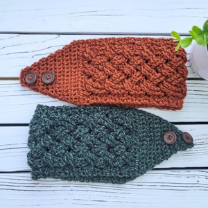 Puede incluir: Dos diademas de ganchillo hechas a mano. Una es de color óxido cálido y la otra es de color verde azulado intenso. Ambas diademas presentan un diseño de punto trenzado texturizado y están adornadas con dos botones de madera marrón en un lado. Las diademas se muestran sobre una superficie de madera blanca.