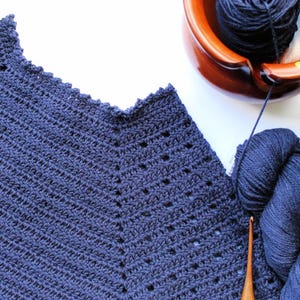 May include: A close-up of a navy blue crocheted garment with a scalloped neckline. The garment is made with a stitch pattern that creates a diagonal texture. Two balls of navy blue yarn are visible in the background.