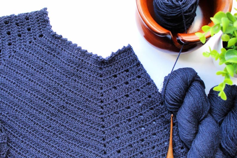 May include: A close-up of a navy blue crocheted garment with a scalloped neckline. The garment is made with a stitch pattern that creates a diagonal texture. Two balls of navy blue yarn are visible in the background.