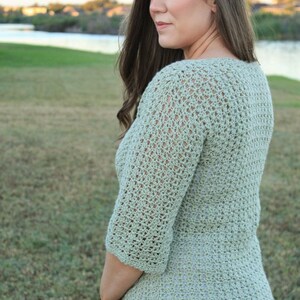 May include: A light green crochet top with three-quarter sleeves. The top has a textured, open weave design. The person is wearing blue jeans and a white undershirt. The background shows a grassy area and a body of water.