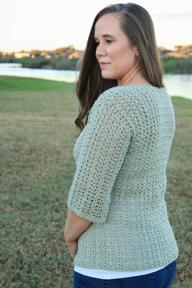 May include: A light green crochet top with three-quarter sleeves. The top has a textured, open weave design. The person is wearing blue jeans and a white undershirt. The background shows a grassy area and a body of water.