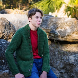 May include: A young person wearing a green crocheted cardigan over a red t-shirt and blue jeans. The person is sitting on a rock with a rocky background.