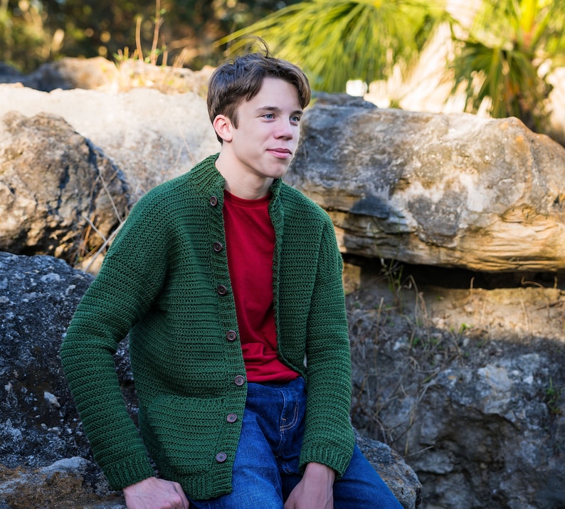 May include: A young person wearing a green crocheted cardigan over a red t-shirt and blue jeans. The person is sitting on a rock with a rocky background.