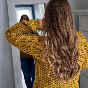 May include: A woman wearing a yellow waffle knit sweater with long, wavy brown hair. She is looking at her reflection in a mirror.