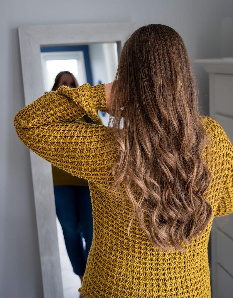 May include: A woman wearing a yellow waffle knit sweater with long, wavy brown hair. She is looking at her reflection in a mirror.
