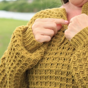 May include: A close-up of a mustard yellow crocheted sweater with a textured stitch pattern. The sweater has a V-neck and long sleeves.