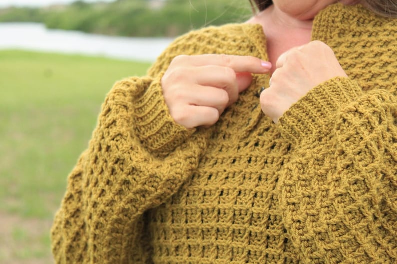 May include: A close-up of a mustard yellow crocheted sweater with a textured stitch pattern. The sweater has a V-neck and long sleeves.