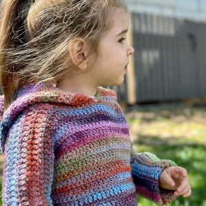 May include: A young child wearing a colorful striped crocheted hooded sweater. The sweater has a variety of colors including pink, purple, blue, green, and orange.