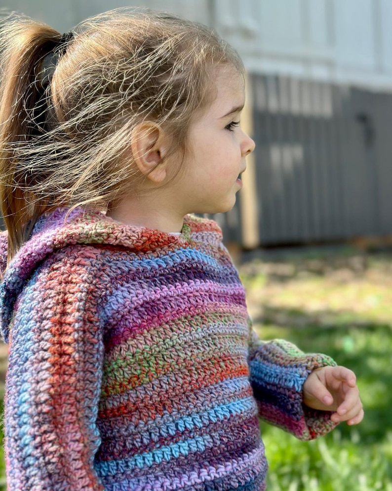 May include: A young child wearing a colorful striped crocheted hooded sweater. The sweater has a variety of colors including pink, purple, blue, green, and orange.