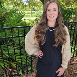 May include: A woman wearing a beige knitted cardigan with long sleeves and a black dress. She is standing in front of a black metal fence with green plants behind it.