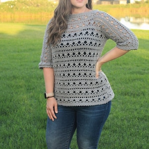 May include: A gray crochet top with a lacy pattern. The top has a boat neck and three-quarter sleeves. The model is wearing the top with blue jeans.