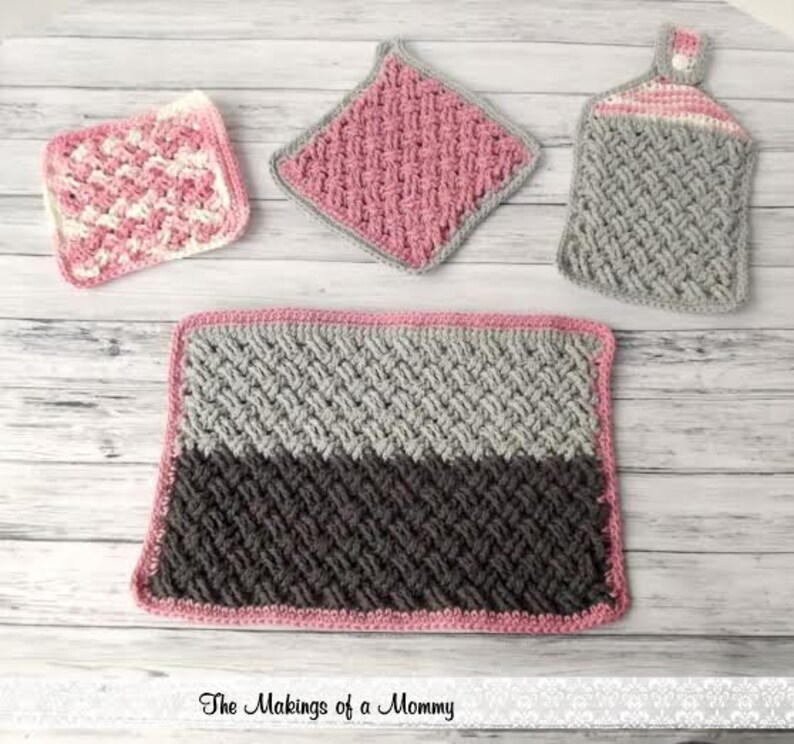 Puede incluir: Una colecci&oacute;n de art&iacute;culos de cocina de ganchillo hechos a mano en tonos rosa, gris y blanco. El conjunto incluye un salvamanteles cuadrado, un pa&ntilde;o de cocina, una toalla para colgar y una alfombra rectangular. Los art&iacute;culos est&aacute;n dispuestos sobre una superficie de madera clara.