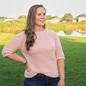 May include: A pink crocheted sweater with a round neckline and three-quarter sleeves. The sweater is made with a simple stitch pattern and has a relaxed fit.