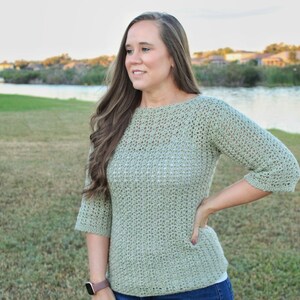 May include: A light green crochet sweater with three-quarter sleeves. The sweater has a round neckline and a textured stitch pattern. The person is wearing a watch on their left wrist. The background includes a lake and green grass.