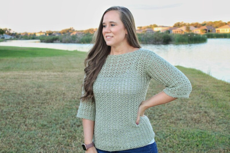 May include: A light green crochet sweater with three-quarter sleeves. The sweater has a round neckline and a textured stitch pattern. The person is wearing a watch on their left wrist. The background includes a lake and green grass.