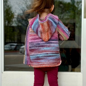 May include: A child wearing a multi-colored striped crocheted hooded sweater with long sleeves and maroon leggings. The child is standing in front of a glass door.