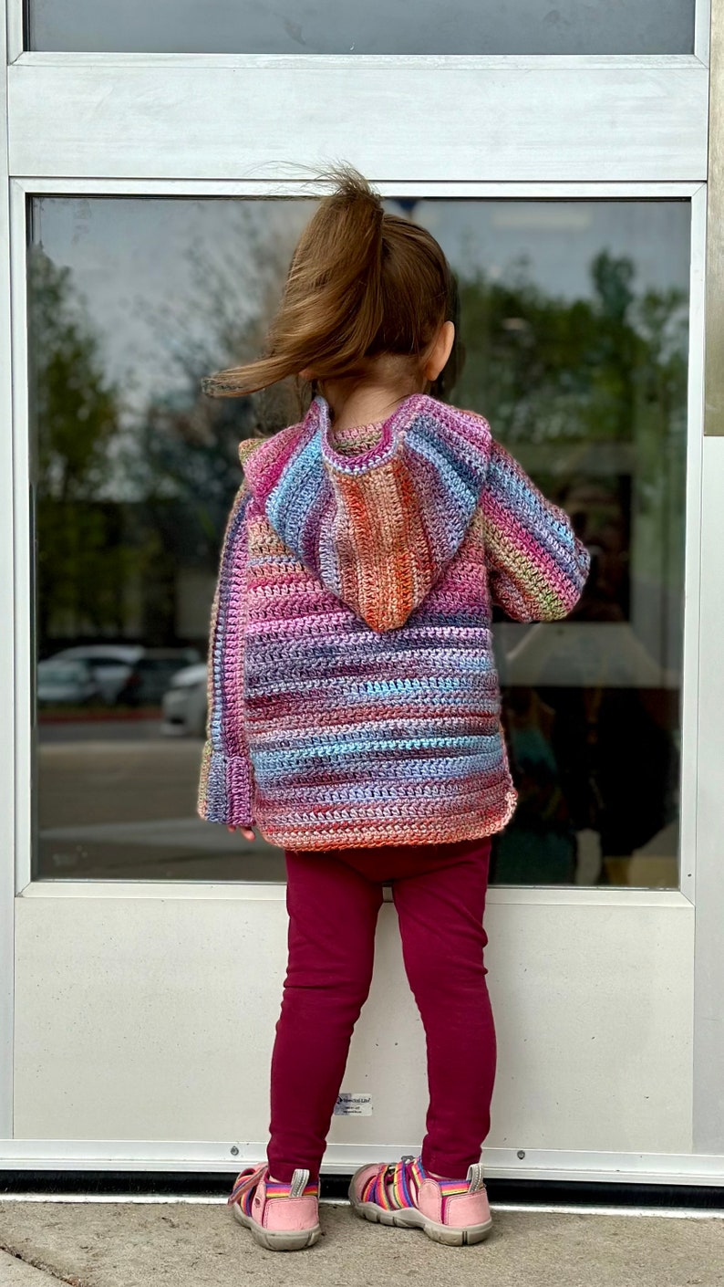 May include: A child wearing a multi-colored striped crocheted hooded sweater with long sleeves and maroon leggings. The child is standing in front of a glass door.