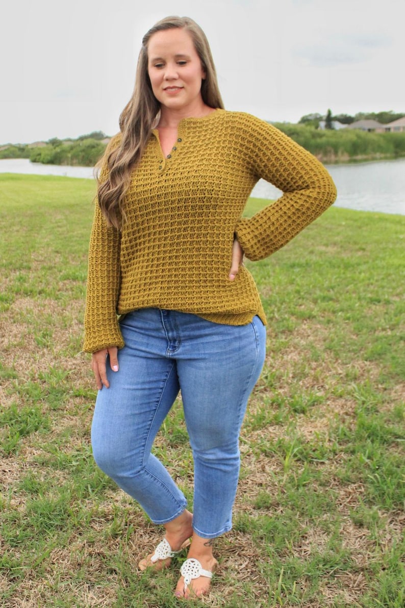 May include: A woman wearing a mustard yellow waffle knit sweater with a button closure at the neck and blue jeans.
