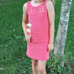 May include: A young girl wearing a pink crocheted dress and gray sandals, holding a bouquet of white daisies. She is standing in a grassy area next to a birch tree.