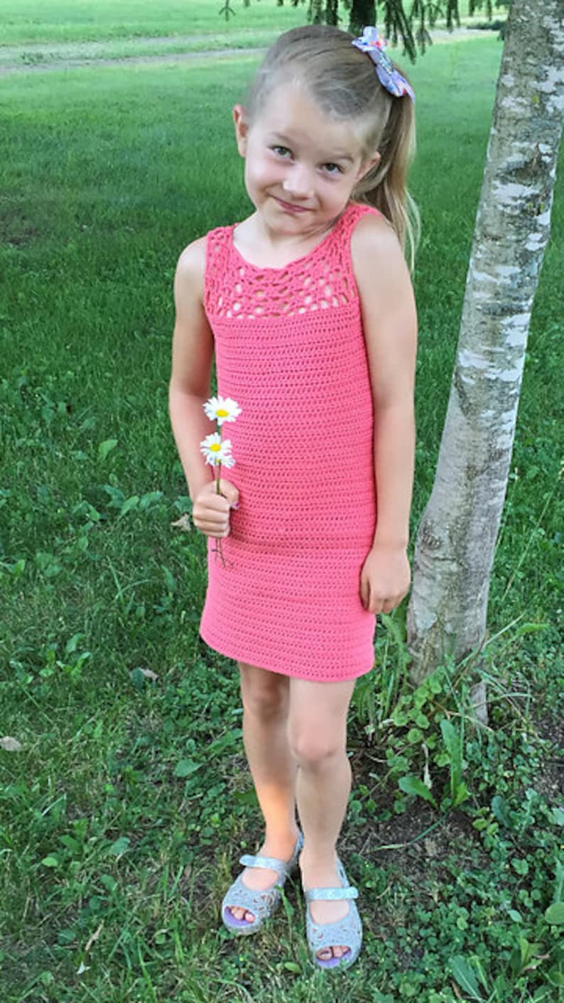 May include: A young girl wearing a pink crocheted dress and gray sandals, holding a bouquet of white daisies. She is standing in a grassy area next to a birch tree.