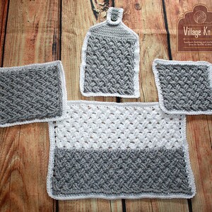 Puede incluir: Un juego de cocina de ganchillo hecho a mano en gris y blanco. El juego incluye un pa&ntilde;o de cocina, dos agarraderas y una alfombra rectangular. Los art&iacute;culos tienen un patr&oacute;n texturizado y ribete blanco. El logotipo de Village Knits est&aacute; en la esquina superior derecha.