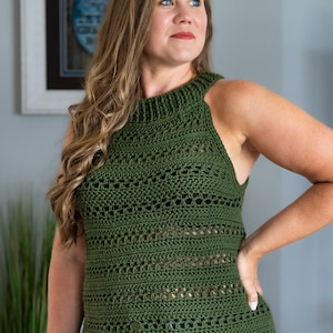 May include: An olive green crochet top with a high neckline and sleeveless design. The top has a textured pattern and a slightly flared hemline. The woman is wearing blue jeans and a watch.