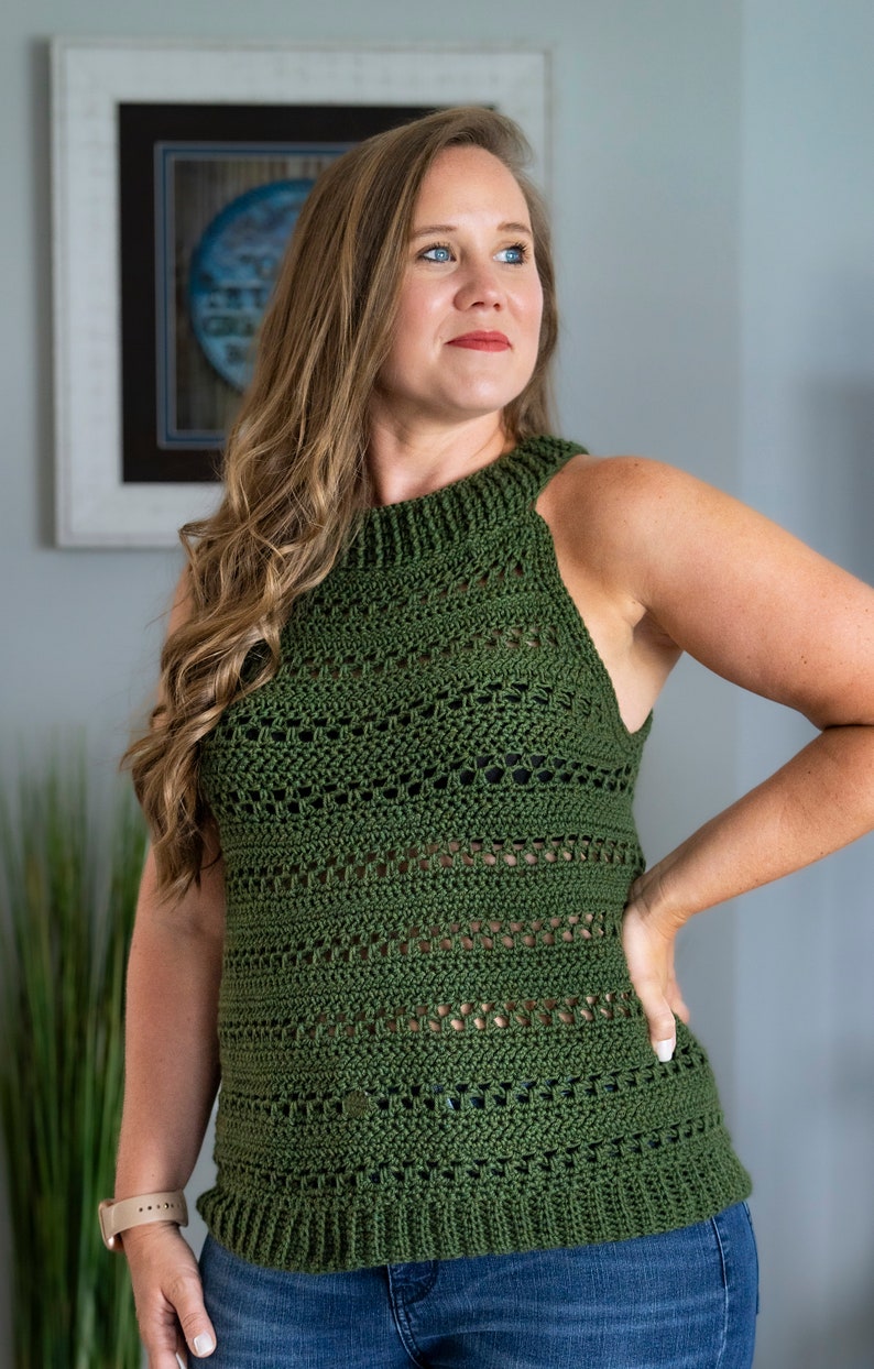 May include: An olive green crochet top with a high neckline and sleeveless design. The top has a textured pattern and a slightly flared hemline. The woman is wearing blue jeans and a watch.