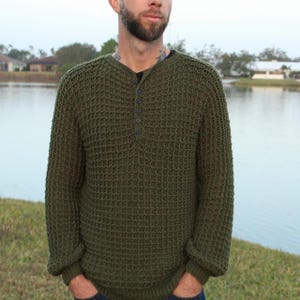 May include: Olive green waffle-knit sweater with a Henley neckline and three dark buttons. The long-sleeved sweater has a textured pattern and a relaxed fit. The model is wearing the sweater with their hands in their pockets.
