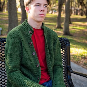 May include: A young person wearing a green knitted cardigan with wooden buttons over a red t-shirt and blue jeans, sitting on a black metal bench.