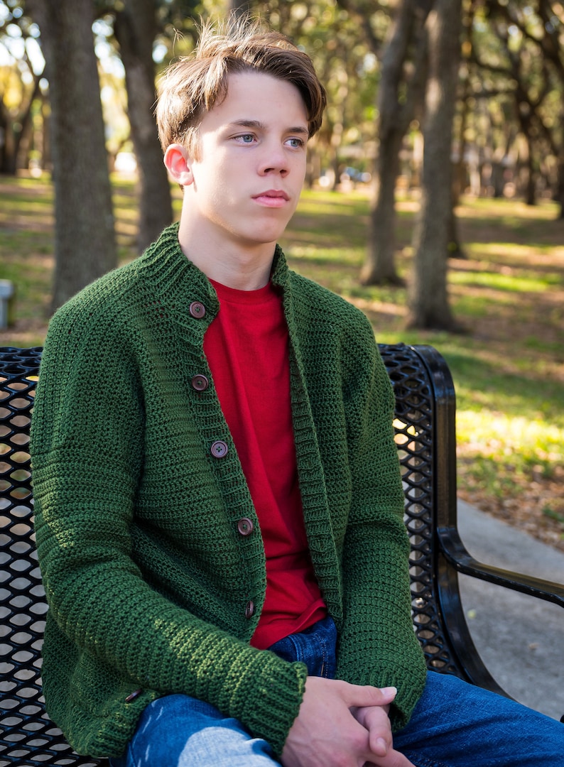 May include: A young person wearing a green knitted cardigan with wooden buttons over a red t-shirt and blue jeans, sitting on a black metal bench.