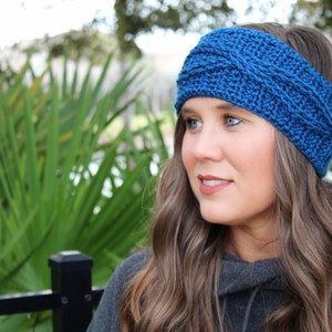 May include: A blue knitted headband with a cable knit pattern. The headband is worn by a person with long brown hair.