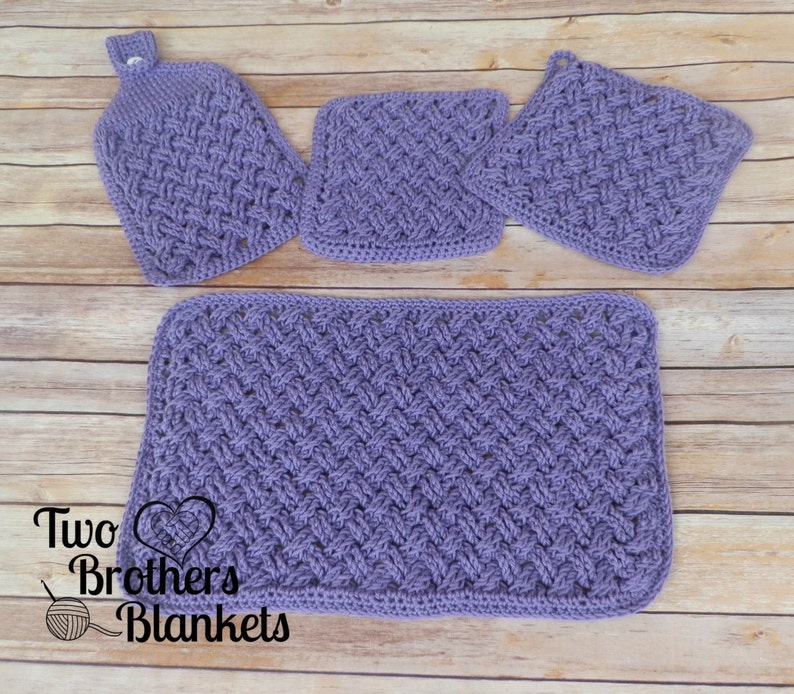 Puede incluir: Un juego de cuatro art&iacute;culos de cocina de ganchillo de color lavanda: una toalla colgante, dos agarraderas cuadradas y un pa&ntilde;o de cocina rectangular. Los art&iacute;culos presentan un patr&oacute;n tejido texturizado. El logotipo "Two Brothers Blankets" es visible en la esquina inferior izquierda.