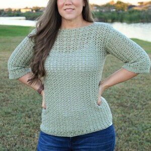 May include: A light green crochet sweater with three-quarter sleeves and a round neckline. The sweater is paired with blue jeans. The person is standing outside on a grassy area.