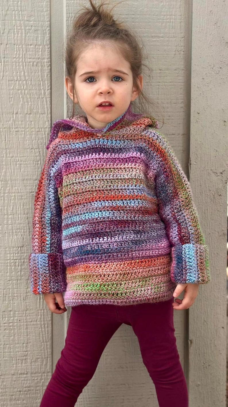 May include: A young child wearing a colorful striped crocheted hooded sweater with long sleeves and burgundy leggings.