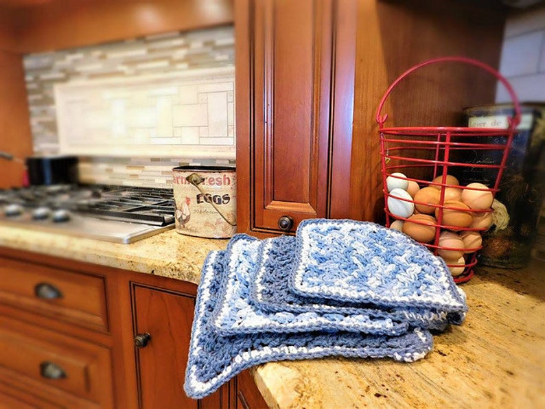 Puede incluir: Una pila de pa&ntilde;os de cocina cuadrados de ganchillo hechos a mano en tonos azules y blancos se encuentra sobre una encimera de cocina. Una cesta de alambre roja llena de huevos marrones est&aacute; en el fondo, junto con un recipiente vintage para huevos.