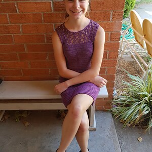 May include: A young person wearing a purple crochet dress with a lacy neckline. They are sitting on a wooden bench with their legs crossed and wearing black shoes.