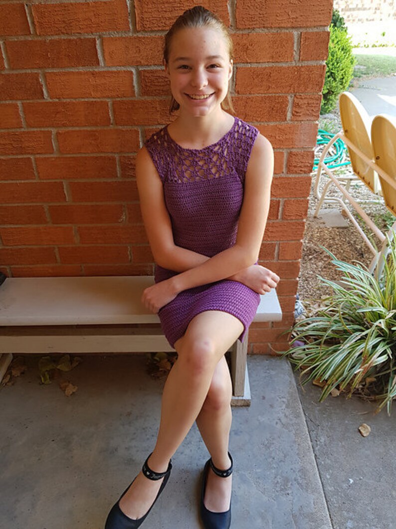May include: A young person wearing a purple crochet dress with a lacy neckline. They are sitting on a wooden bench with their legs crossed and wearing black shoes.