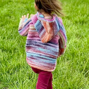 May include: A child wearing a colorful striped crocheted hooded sweater with a long-sleeved design. The sweater has a variety of colors, including pink, blue, purple, and orange. The child is wearing maroon pants and is walking on a grassy field.