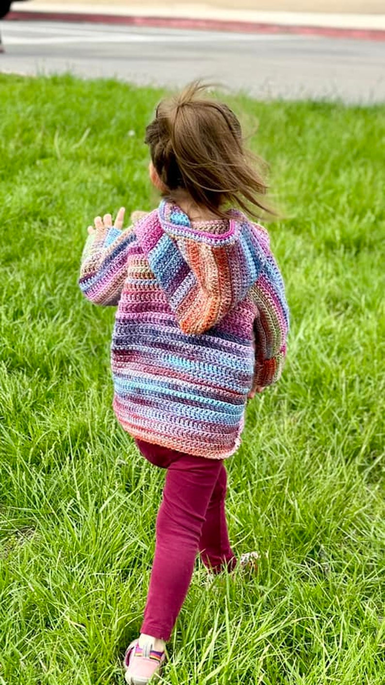 May include: A child wearing a colorful striped crocheted hooded sweater with a long-sleeved design. The sweater has a variety of colors, including pink, blue, purple, and orange. The child is wearing maroon pants and is walking on a grassy field.