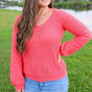 May include: Coral-colored, long-sleeved sweater with a V-neck. The sweater has a ribbed texture and is paired with blue jeans. The background features green grass and a body of water.