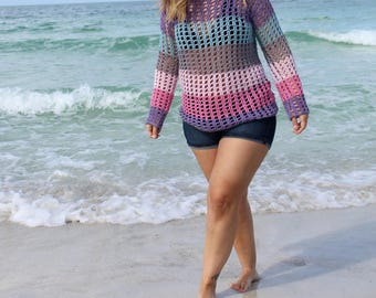 May include: A woman wearing a multi-colored crochet top with a loose fit and long sleeves. The top is striped with shades of pink, purple, blue, and white. The woman is standing on a sandy beach with ocean waves in the background.