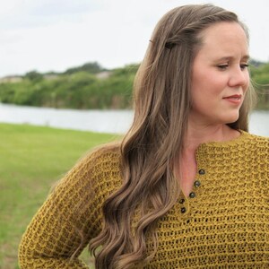 May include: A woman with long brown hair wearing a mustard yellow crocheted sweater with a button detail at the neckline. She is standing in front of a body of water and green grass.