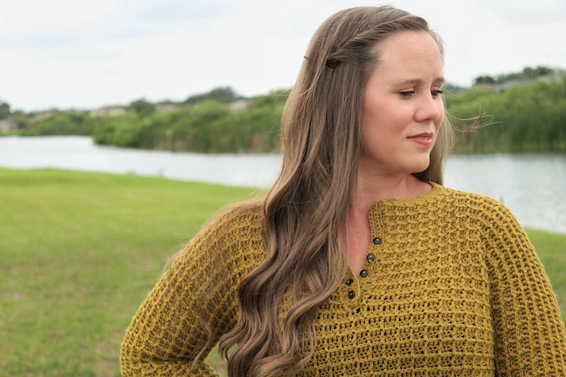 May include: A woman with long brown hair wearing a mustard yellow crocheted sweater with a button detail at the neckline. She is standing in front of a body of water and green grass.