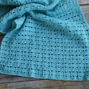 May include: A crocheted scarf in a light turquoise color. The scarf has an open, lacy design with a repeating pattern of small, fan-shaped elements. The scarf is draped over a wooden surface.