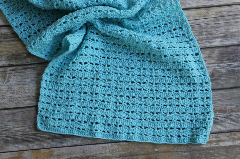 May include: A crocheted scarf in a light turquoise color. The scarf has an open, lacy design with a repeating pattern of small, fan-shaped elements. The scarf is draped over a wooden surface.