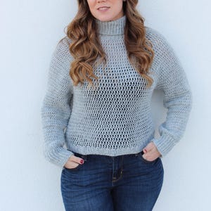 May include: A light gray crocheted sweater with a turtleneck and a loose fit. The sweater has a delicate open stitch pattern.