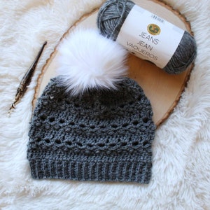 May include: A gray crocheted beanie with a white faux fur pom pom. The beanie is made with a lacy stitch pattern. The yarn is labeled "Jeans Jean Vaqueros".